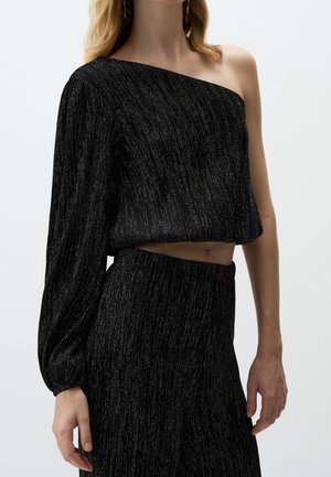 Kobieta ubrana w czarny, asymetryczny crop top z długim rękawem i pasujące, wysokie spodnie z delikatnymi srebrnymi pionowymi paskami.