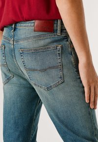 Blå denimjeans med en tettsittende passform, synlige overlocksting og en brun lærlogo på bakre linning.
