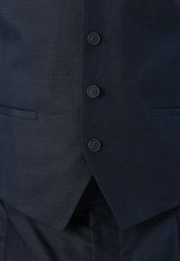 Dunkelmarineblauer Anzugweste mit drei geschlossenen Knöpfen, zwei vorderen Paspeltaschen und dazu passenden Anzughosen, die darunter sichtbar sind.