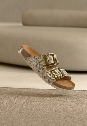 Beige snake-patterned sandal with two gold buckle straps and cork footbed floating above a beige carpet in a neutral room setting.