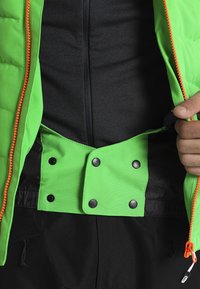 Personne attachant une veste vert vif avec une fermeture éclair orange par-dessus des vêtements noirs, mettant l'accent sur la fermeture à boutons-pression et les détails de la fermeture éclair.