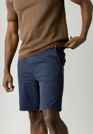 Mężczyzna w brązowej bluzce z krótkim rękawem i granatowych szortach, lekko ciągnący dolną krawędź bluzki na zewnątrz prawą ręką.