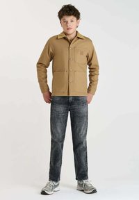 Beige fleece jack met een kraag, twee voorzakken, drukknoopsluiting, gestructureerde stof, gecombineerd met donkere spijkerbroek en grijze sneakers.
