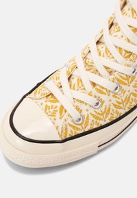 Baskets en toile jaune et blanche avec motif de feuilles, lacets blancs, embout crème et bordure noire sur semelle blanche.