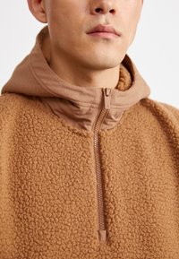 Pullover de fleece castanho com capuz liso, apresentando um fecho de correr à frente e um tecido texturado. Marcação discreta no puxador do fecho.