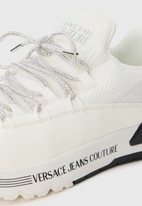 Sneaker bianco di Versace Jeans Couture realizzato in tessuto testurizzato, con lacci decorati e logo stampato di lato sulla suola in lettere nere.
