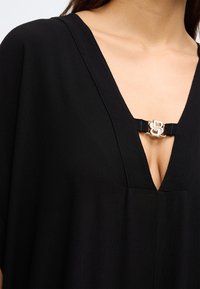 Fekete ruhadarab mély V-nyakkal, texturált anyaggal és középen ezüst színű díszítéssel, amelyet egy kontrasztos pánt emel ki.