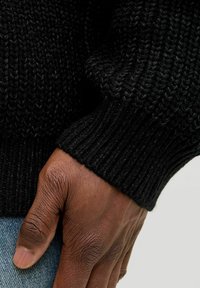 Eine dunkelhäutige Hand liegt in der Nähe von blauen Jeans und trägt einen schwarzen, grob gestrickten Pulloverärmel mit sichtbarem Rippenmuster.