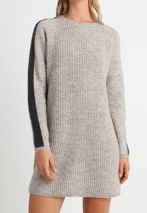 Vrouw draagt een gebreide trui-jurk met lange mouwen tot op de knie in lichtgrijs met een donkergrijze streep langs de buitenkant van de mouwen.