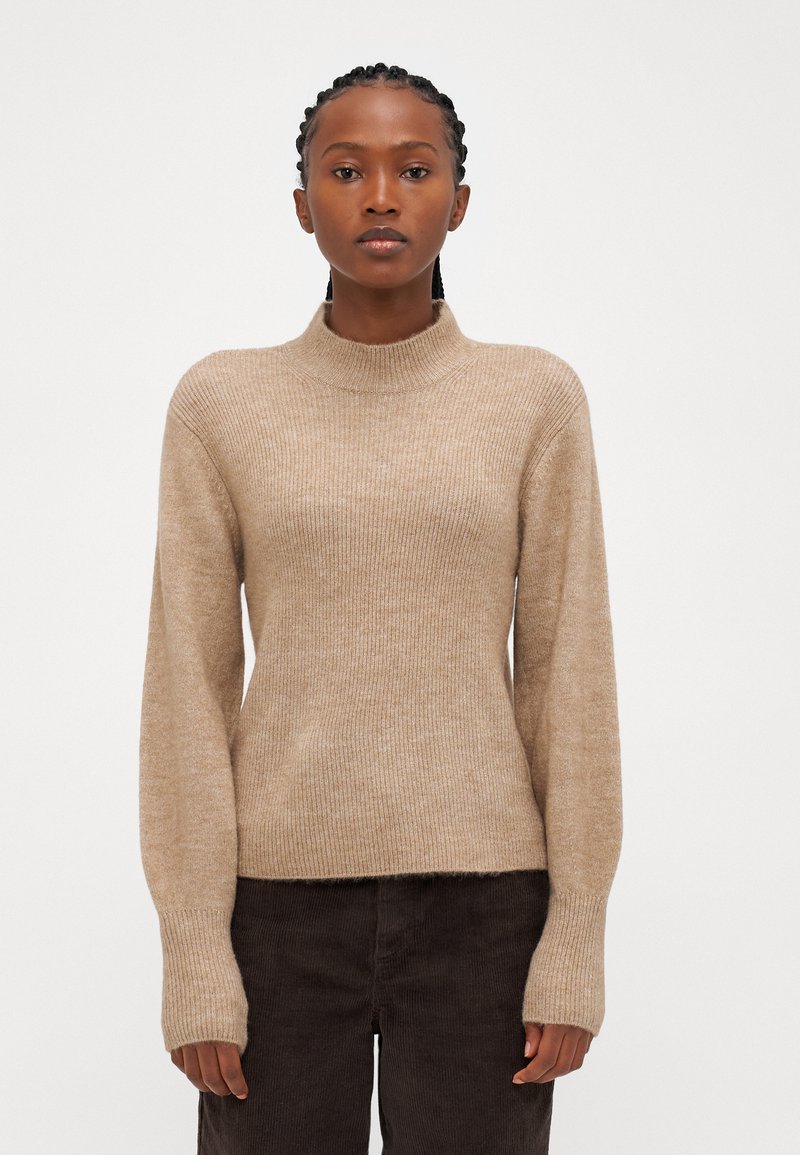 Maglione beige lavorato a maglia con collo alto, texture a coste e maniche lunghe, abbinato a pantaloni in corduroy scuri.
