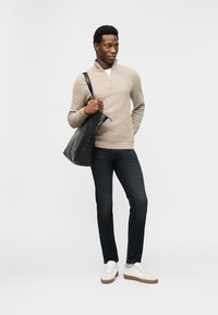 Νέος άνδρας που φοράει μπεζ πουλόβερ με φερμουάρ έως τη μέση, σκούρο τζιν, λευκά αθλητικά παπούτσια, κρατώντας μαύρη δερμάτινη τσάντα ώμου, στέκεται μπροστά σε απλό φόντο.