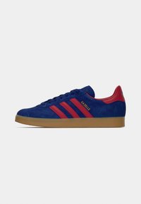 Sapatilha Adidas Gazelle em camurça azul com riscas vermelhas, aba do calcanhar vermelha, sola de borracha e texto dourado "GAZELLE" na lateral.