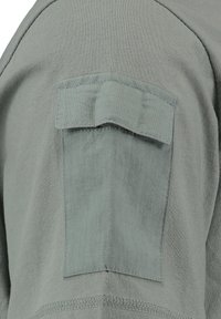 Zielona materiałowa rękawica z haftowaną kieszenią na klapę, charakteryzującą się prostokątnym kształtem i widocznymi detalami szwów.