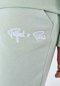 Joggers de algodón verde claro con una textura suave, con texto bordado en blanco "Project + Paris" en la zona del bolsillo frontal.