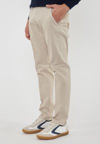 Pantaloni in cotone beige con vestibilità dritta, tasche laterali e una leggerissima trama, abbinati a sneakers bianche con dettagli blu navy e suole in gomma.