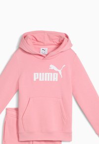 Rožinė gobtuvė pagaminta iš medvilnės ir sintetinio pluošto mišinio, su priekinėmis kišenėmis, reguliuojamu raišteliu ir balta Puma logotipu ant krūtinės.