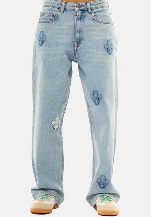 Vaqueros sueltos azul claro con parches de flor de lis combinados con zapatillas beige con cordones verdes y blancos.