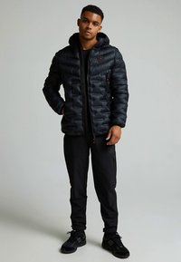 Svart dunjakke med hette, med glidelås og synlige sømmer. Kombinert med svarte bukser og svarte joggesko.