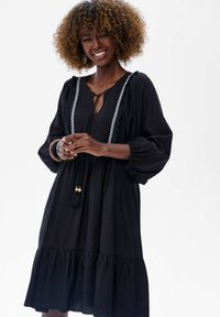 Vrouw met krullend haar die glimlacht, gekleed in een zwarte jurk met pofmouwen, kantafwerking, en houten armbanden om haar pols.