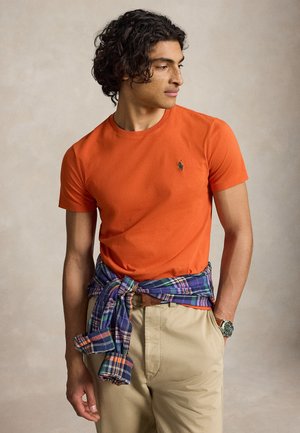 Ung mand med krøllet hår iført en orange kortærmet T-shirt, beige bukser og en blå ternet skjorte bundet rundt om livet.