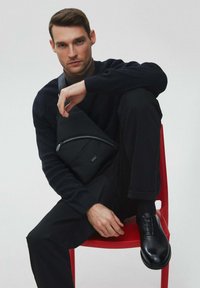 Czarna tkaninowa torba na ramię z przednim zamkiem, noszona przez osobę w czarnym swetrze i spodniach, siedzącą na czerwonym krześle. Gładkie skórzane buty.