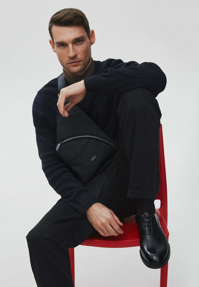 Czarna tkaninowa torba na ramię z przednim zamkiem, noszona przez osobę w czarnym swetrze i spodniach, siedzącą na czerwonym krześle. Gładkie skórzane buty.