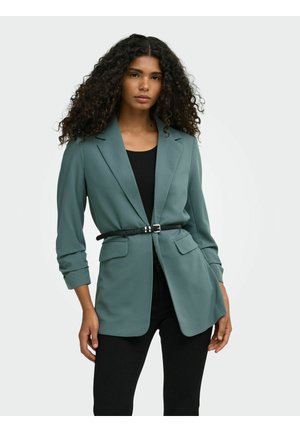 Donna con capelli ricci che indossa una giacca color teal stretta in vita con una cintura nera, top nero e pantaloni neri su uno sfondo semplice.