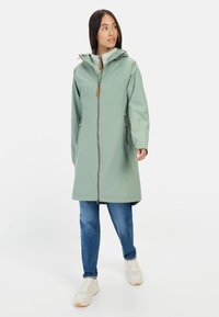Lichtgroene waterdichte jas met een capuchon, voorkant met rits en zijzakken. Gepaard met blauwe spijkerbroeken en witte sneakers.