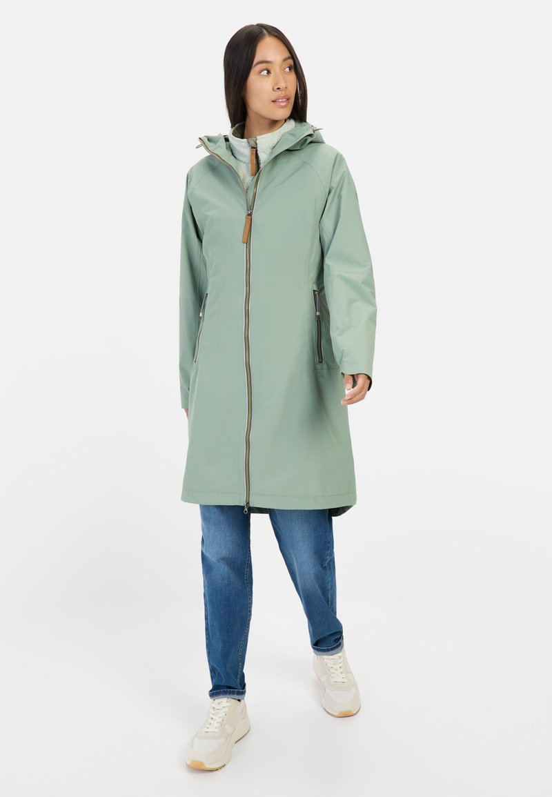Lichtgroene waterdichte jas met een capuchon, voorkant met rits en zijzakken. Gepaard met blauwe spijkerbroeken en witte sneakers.