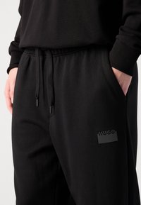 Zwarte sweatpants van zacht materiaal, met een elastische tailleband met trekkoord, zijzakken en een zwart logo op de dij.