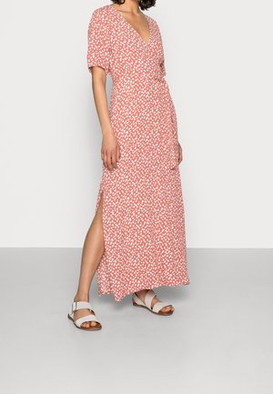 Maxi-jurk in koraal met wit loofpatroon, V-hals, korte mouwen en een zijsplits. Gecombineerd met lichtgekleurde sandalen.