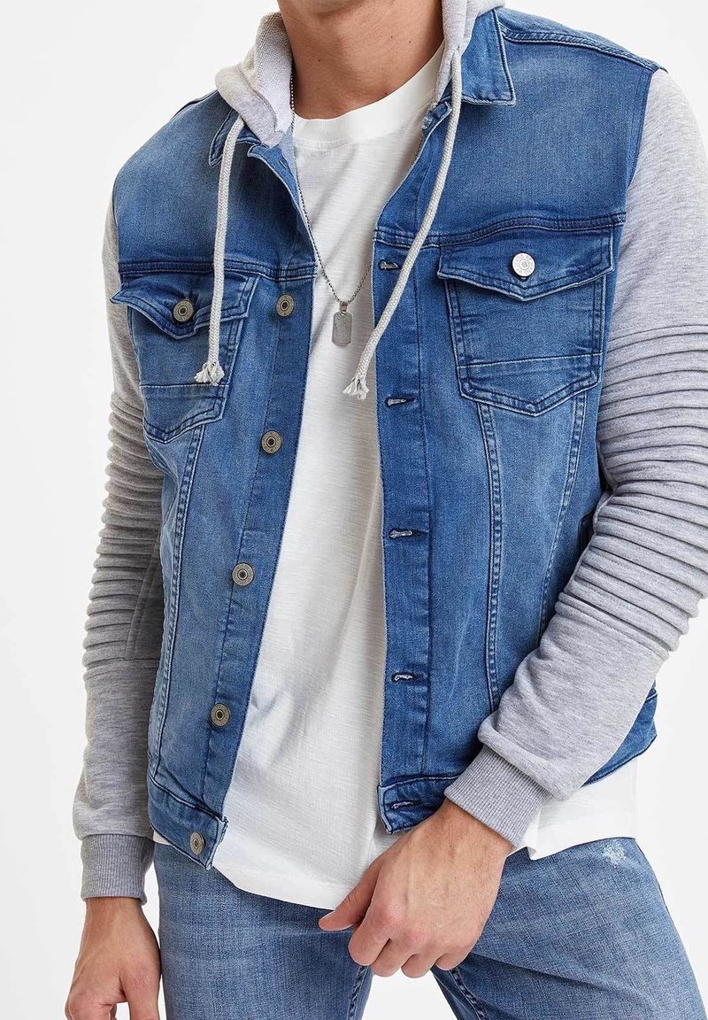 Homme portant un gilet en jean bleu avec des manches de sweat à capuche gris, un t-shirt blanc, un jean bleu et un collier avec une plaque de chien argentée.
