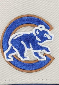 Circular patch features a blue bear design embroidered in white, outlined with brown. Background is light beige with textured fabric.