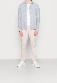 Chemise à rayures bleues et blanches boutonnée, associée à un pantalon beige clair et des baskets blanches. Le modèle se tient debout, les mains le long du corps.