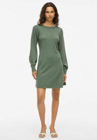 Donna che indossa un abito verde oliva lungo fino al ginocchio, con maniche lunghe a sbuffo e scarpe beige con punta aperta, in piedi di fronte a uno sfondo bianco.