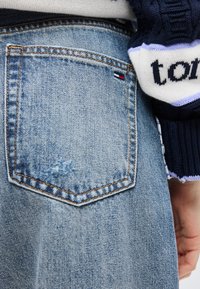 Τζιν παντελόνι με πίσω τσέπη που φέρει μια μικρή ετικέτα με κόκκινη, λευκή και μπλε σημαία, φορεμένο με έναν μπλε και λευκό μανίκι πουλόβερ ορατό.