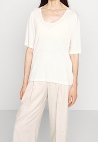 Bílý krátký rukáv, top s véčkovým výstřihem, vyrobený z hladkého materiálu. V kombinaci s béžovými volnými kalhotami s texturovaným vzorem.