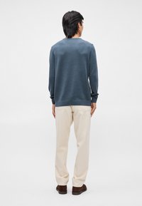 Blå strikkegenser med lange ermer, rund hals, strukturert mønster. Kombinert med kremfargede bukser og brune sko. Bakperspektiv.