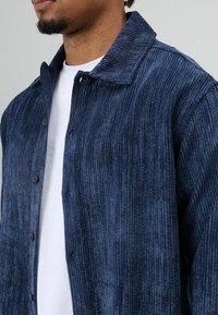 Marineblauw corduroy shirt met verticale ribbeltjesstructuur, voorzien van een knoopsluiting aan de voorkant en een kraag, gedragen over een wit T-shirt.