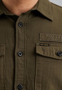 Camicia verde oliva in tessuto strutturato, con bottoni neri, una tasca sul petto con etichetta di branding e colletto a punta.