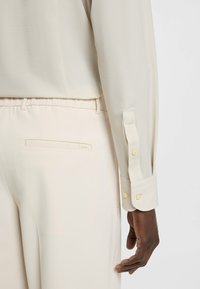 Camicia beige a maniche lunghe con una leggera trama e polsini con bottoni, abbinata a pantaloni crema con tasca posteriore e un sottile dettaglio di branding.