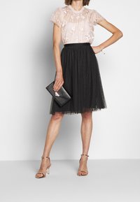 Blouse rose clair brodée avec manches à volants, associée à une jupe en tulle noire et des talons dorés à brides. Pochette noire en forme d'enveloppe.