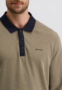 Beige strukturiertes Poloshirt mit einem dunkelblauen Kragen, einer drei-knöpfigen Knopfleiste und subtiler Markenkennzeichnung auf der Brust.