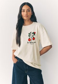 Krém színű, rövid ujjú pamut póló, piros paradicsomok és zöld levelek grafikájával, "la dolce vita Napoli" felirattal.