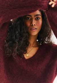 Bordowy, oversize sweter wykonany z miękkiej, teksturowanej tkaniny z dekoltem w serek. Posiada długie rękawy oraz delikatne wykończenia dzianinowe przy krawędziach.