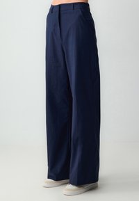 Pantalons larges bleu marine en tissu léger. Comprend des poches avant et une texture lisse, associés à des chaussures de couleur claire.