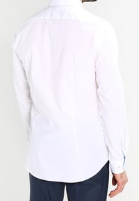 Biała koszula z długim rękawem, kołnierzykiem z zapięciem na guziki, gładką fakturą, zaokrąglonym dołem oraz kontrastującym niebieskim akcentem wewnątrz kołnierzyka i mankietów.