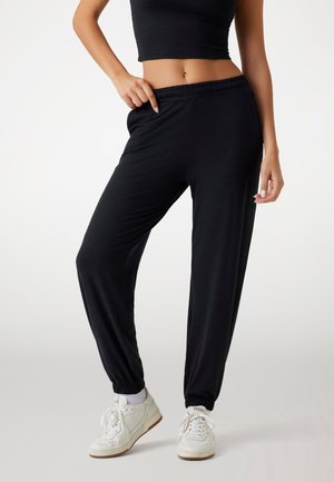 Kobieta ubrana w czarne spodnie dresowe, białe skarpetki i białe trampki, z jedną ręką na pasku spodni, stojąca na jednolitym tle.