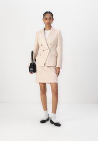 Fato de tweed rosa claro com blazer de duplo botão e saia justa, apresentando botões dourados e barra desfiada. Mala preta e sapatos rasos.