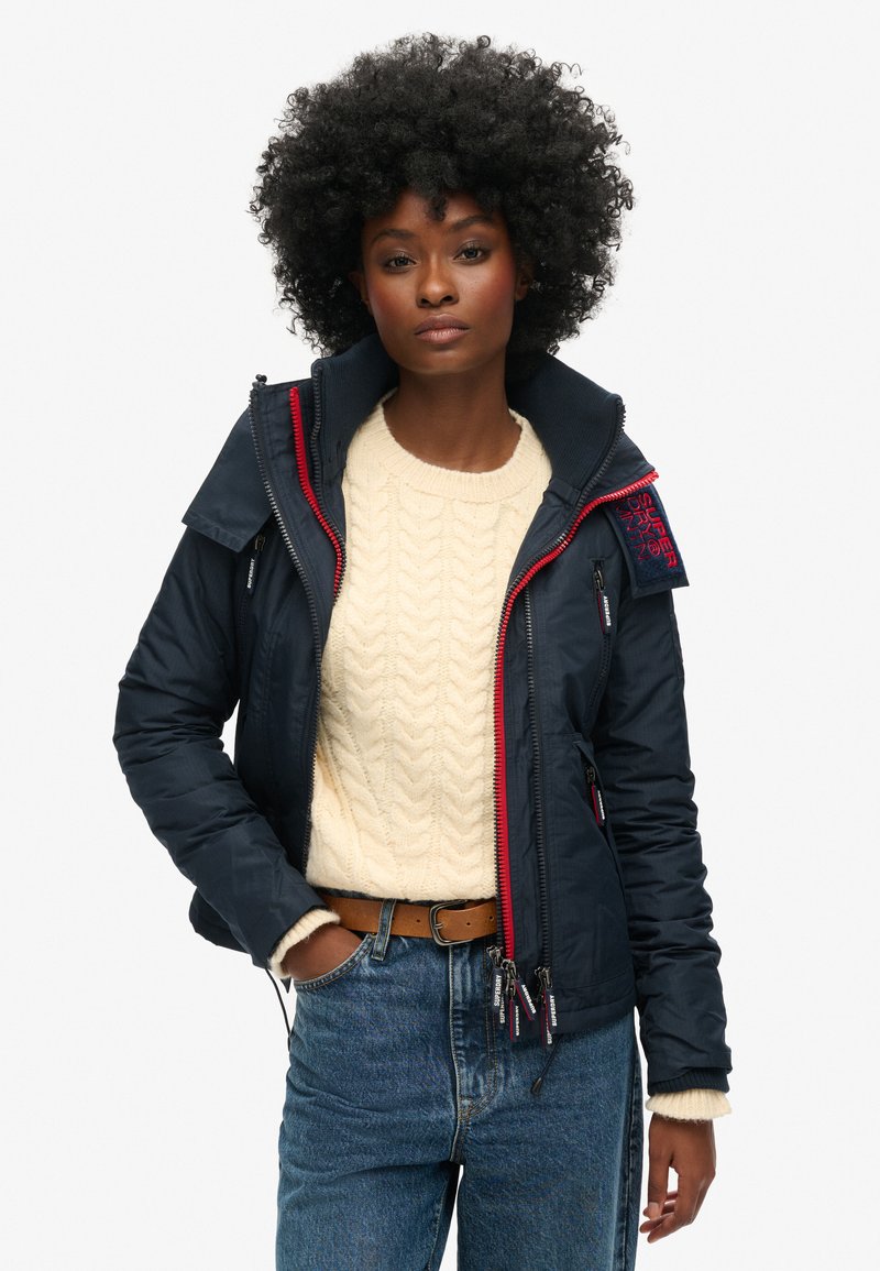 Marineblauwe pufferjack met een hoge kraag, rode ritsaccenten en meerdere zakken, gedragen over een crèmekleurige kabelgebreide trui en blauwe jeans.